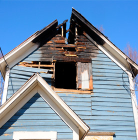 Damaged blue house with charred roof and exposed interior from fire damage, highlighting the need for public insurance adjusting services.